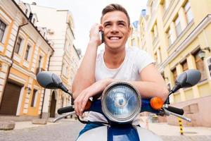 Jung und frei. Fröhlicher junger Mann sitzt auf Roller und telefoniert mit dem Handy foto