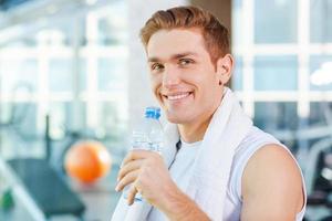 hydratisiert bleiben. hübscher junger Mann, der ein Handtuch auf den Schultern trägt und Wasser trinkt, während er im Fitnessstudio steht foto