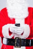 Nachricht an Elfen. Nahaufnahme des Weihnachtsmanns, der Handy hält, während er vor grauem Hintergrund steht foto