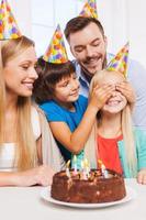 Überraschen Sie die glückliche vierköpfige Familie, die den Geburtstag eines glücklichen kleinen Mädchens feiert, das am Tisch sitzt, während ihr Bruder ihre Augen mit den Händen bedeckt und lächelt foto