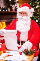 überprüft seine Weihnachtsliste. Konzentrierter Weihnachtsmann, der auf seinem Stuhl sitzt und einen Brief liest, während Umschläge auf dem Tisch liegen foto