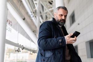 Reifer Mann, der darauf wartet, am Flughafen in einen Flug einzusteigen und auf sein Handy zu schauen foto