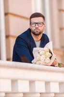 wartet auf seine Freundin. hübscher junger Mann in schicker Jacke, der einen Blumenstrauß hält und wegschaut, während er im Freien steht foto