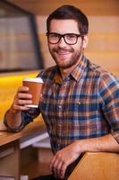 Heißen und frischen Kaffee genießen. glücklicher junger mann, der kaffeetasse hält und lächelt, während er im café sitzt foto