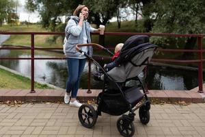 junge fürsorgliche mutter mit einer tasse kaffee in den händen neben dem kinderwagen mit ihrem neugeborenen kind, während sie im park spazieren geht, spricht mit einem lächeln im gesicht am telefon foto