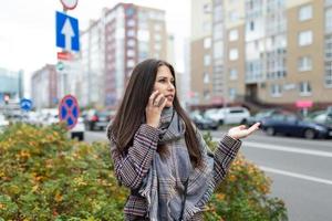 Stilvolle junge Frau spricht auf einem Handy in der Nähe der Straße mit Gesten, zeigt Verwirrung, fängt ein Taxi foto