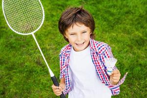 kleiner Weltmeister. Blick von oben auf den glücklichen kleinen Jungen, der Badmintonschläger und Federball hält, während er auf grünem Gras steht foto