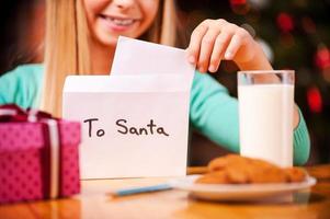 Brief an den Weihnachtsmann. Nahaufnahme eines fröhlichen kleinen Mädchens, das einen Brief an den Weihnachtsmann in den Umschlag steckt, während es mit einem Glas Milch und Keksen darauf am Tisch sitzt foto