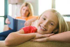 Zeit in der Nähe der Mutter genießen. glückliches kleines Mädchen, das auf der Couch sitzt und lächelt, während ihre Mutter im Hintergrund ein Buch liest foto