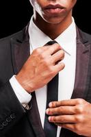 perfekt bis ins letzte Detail. Modischer junger afroamerikanischer Mann, der seine Krawatte anpasst, während er vor schwarzem Hintergrund steht foto