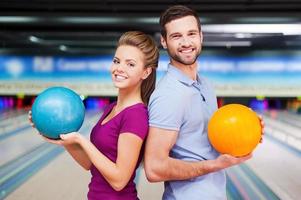 wir sind ein Team. Fröhliche junge Männer und Frauen, die über die Schultern schauen und Bowlingkugeln halten, während sie gegen Bowlingbahnen stehen foto