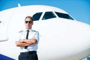 bereit für den Himmel. Selbstbewusster männlicher Pilot in Uniform, der die Arme verschränkt hält, während er in der Nähe des Flugzeugs steht foto