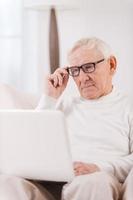 selbstbewusster Heimarbeiter. Schöner älterer Mann, der am Laptop arbeitet, während er in seiner Wohnung auf einem Stuhl sitzt foto