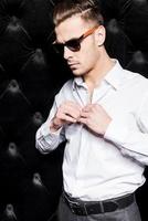 trägt sein Lieblingsshirt. hübscher junger Mann mit Sonnenbrille, der sein weißes Hemd zuknöpft, während er vor schwarzem Hintergrund steht foto