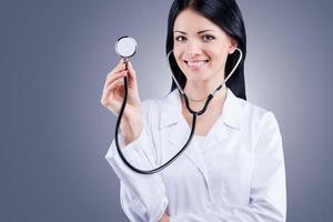 Atmen Sie, bitte zuversichtlich, Ärztin in weißer Uniform, die Stethoskop hält und lächelt, während sie vor grauem Hintergrund steht foto