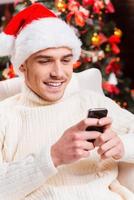 Nachricht an seinen Nächsten schreiben. Gutaussehender junger Mann mit Weihnachtsmütze SMS auf seinem Handy und lächelnd mit Weihnachtsbaum im Hintergrund foto