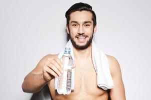 Trinken Sie etwas Wasser Junger muskulöser Mann, der eine Flasche mit Wasser in der Hand ausstreckt und lächelt, während er vor grauem Hintergrund steht foto