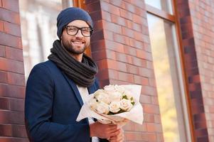 Auf sie warten. hübscher junger Mann in eleganter Freizeitkleidung, der einen Blumenstrauß hält und lächelt, während er auf der Straße steht foto