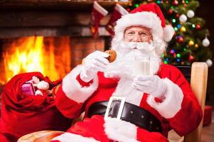 mein lieblingsessen fröhlicher weihnachtsmann, der ein glas mit milch und keks hält, während er an seinem stuhl mit kamin und weihnachtsbaum im hintergrund sitzt foto