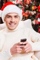 SMS an seinen Nächsten. Gutaussehender junger Mann mit Weihnachtsmütze SMS auf seinem Handy und lächelnd mit Weihnachtsbaum im Hintergrund foto