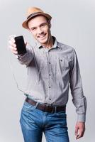 Dies ist mein Lieblingslied. Schöner junger Mann in Fedora, der seinen MP3-Player zeigt und lächelt, während er vor grauem Hintergrund steht foto