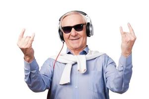 für immer jung. Fröhlicher älterer Mann mit Kopfhörern, der Musik hört und Handzeichen zeigt, während er vor weißem Hintergrund steht foto