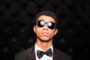 Einfach der beste modische junge afroamerikanische Mann, der eine Sonnenbrille trägt, während er vor dunklem Hintergrund steht foto