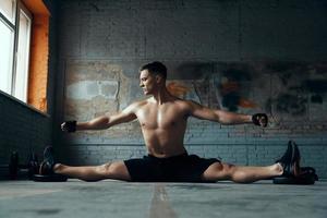Selbstbewusster junger Mann, der den Spagat macht und die Arme ausgestreckt hält, während er im Fitnessstudio trainiert foto