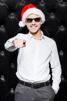 Bist du bereit für eine Party? Schöner junger Mann mit Sonnenbrille und Weihnachtsmütze, der auf dich zeigt und lächelt, während er vor schwarzem Hintergrund steht foto