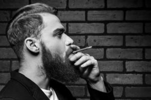 Mann raucht. Schwarz-Weiß-Porträt eines hübschen jungen bärtigen Mannes, der eine Zigarette raucht, während er gegen eine Ziegelwand steht foto