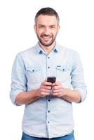 zufrieden mit seinem brandneuen Smartphone. glücklicher junger Mann in Freizeitkleidung hält Smartphone und lächelt, während er isoliert auf weißem Hintergrund steht foto