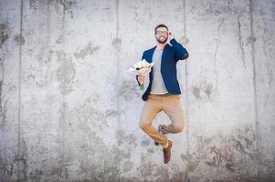 glücklich und verliebt. Fröhlicher junger Mann in schicker Jacke, der einen Blumenstrauß hält und vor die Betonwand springt foto