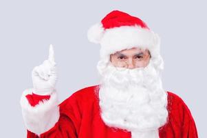Schauen Sie sich diesen traditionellen Weihnachtsmann an, der nach oben zeigt, während er vor grauem Hintergrund steht foto