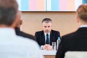 selbstbewusster Business-Trainer. Selbstbewusster reifer Mann in Abendgarderobe, der mit einem großen Monitor auf dem Kopf und mit Menschen im Vordergrund am Tisch sitzt foto