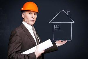 selbstbewusster Architekt. Selbstbewusster älterer Mann in Abendkleidung und Hardhat, der ein mit Kreide gezeichnetes Haus in der Hand hält und eine Blaupause trägt, während er an der Tafel steht foto