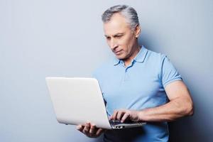 Moderne Technologien immer zur Hand. Fröhlicher älterer Mann in lässigem Laptop, während er vor grauem Hintergrund steht foto