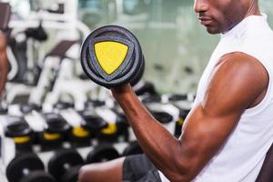 macht seinen Bizeps perfekt. Zugeschnittenes Bild eines jungen afrikanischen Mannes, der im Fitnessstudio mit Hanteln trainiert foto