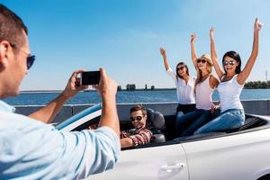 glückliche Momente festhalten. Rückansicht des jungen Mannes, der seine Freunde fotografiert, die im Cabrio sitzen foto