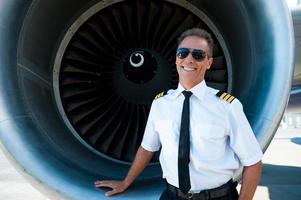 selbstbewusster Pilot. Selbstbewusster männlicher Pilot in Uniform, der sich an das Turbinentriebwerk des Flugzeugs lehnt und lächelt foto