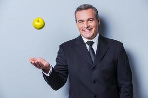 Gesundes Essen für den Erfolg. Selbstbewusster reifer Mann in Abendkleidung, der einen grünen Apfel hochwirft und lächelt, während er vor grauem Hintergrund steht foto