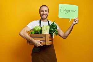 Fröhlicher Mann, der eine Holzkiste mit Gemüse trägt und Bio-Banner vor gelbem Hintergrund zeigt foto
