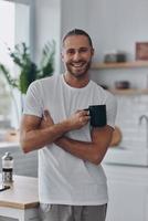 junger Mann, der Kaffee genießt und lächelt, während er in der Küche steht foto