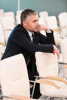 zu langweilig Konferenz. Gelangweilter reifer Mann in Abendkleidung, der die Hand am Kinn hält, während er auf dem Stuhl im leeren Konferenzsaal sitzt foto