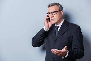 Geschäft besprechen. Selbstbewusster reifer Mann in Abendkleidung, der auf dem Handy spricht und gestikuliert, während er vor grauem Hintergrund steht foto