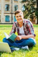 Ich liebe es zu lernen. Fröhlicher männlicher Student, der ein Buch liest und lächelt, während er auf dem Rasen und vor dem Universitätsgebäude sitzt foto