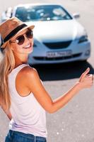 eine Fahrgelegenheit ergattern. schöne junge flippige Frau, die am Straßenrand per Anhalter fährt und mit einem Auto im Hintergrund lächelt foto