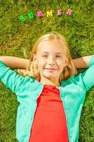 Sommerzeit genießen. Blick von oben auf ein süßes kleines Mädchen, das die Hände hinter dem Kopf hält und lächelt, während es auf dem grünen Gras liegt, mit bunten Plastikbuchstaben, die auf ihrem Kopf liegen foto
