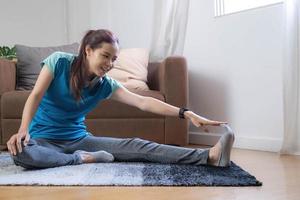Lächelnde asiatische Frau, die zu Hause im Wohnzimmer Yoga-Schulterdehnungs-Online-Kurs macht. Selbstisolation und Training zu Hause während Covid-19. foto