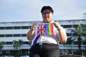 asiatischer junge hält regenbogenflagge und union jack-flagge und trägt krawatte und steht außerhalb des gebäudes, weicher und selektiver fokus, konzept für lgbtq-feier im stolzmonat. foto