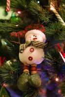 Schneemann aus Filz auf einer Tanne mit Weihnachtsschmuck und verschwommenen Lichtern.